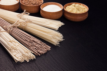 Raw asian noodles with ingredient in wooden bowls on black striped mat background with copy space.