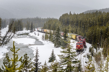 Naklejka premium Gargo Train on Winding Track alongside the Bow River during a Heavy Snowfall. Banff National Park, Alberta, Canada