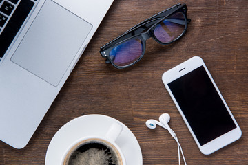 Top view of coffee cup, smartphone with earphones, laptop and glasses on office desk