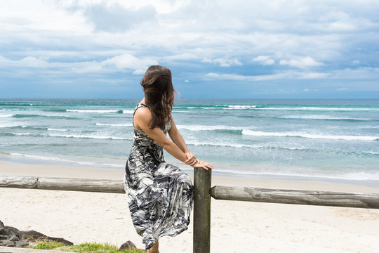 The barefoot woman with long dark hair wearing long romantic dress sitting on the wooden fence and facing on the ocean is looking into the distance on the stormy ocean in Byron Bay, Australia