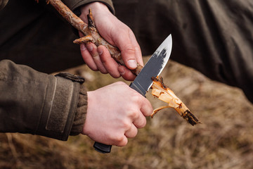 Close-up hand hunter man with knife cut a wooden stick for campfire in the forest.  .