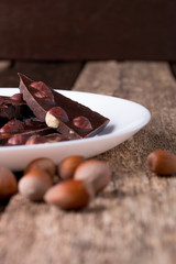 Chocolate pieces with hazelnut nuts on white plate wooden background.
