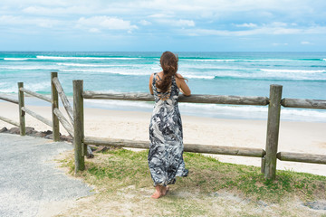 The woman with long dark hair wearing long romantic dress facing on the ocean is looking into the...