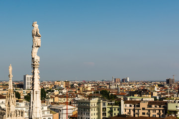 Statues on steeples at the roof of Il Duomo di Milano. Panoramic view on Milano city from the top of the cathedral and antique statue on pillar at background.