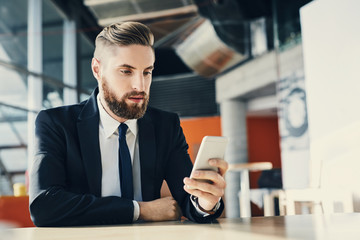 Young man in suit sitting by table browsing his smartphone at modern office