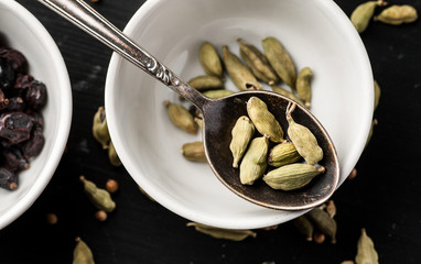 White ceramic bowl with cardamon seeds in it and metal spoon above on a black wooden table, top view