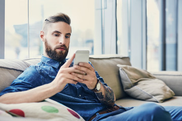 Stylish tottooed man using his smartphone,  sitting on sofa