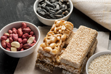 Peanut and sesame brittle stacked on a piece of paper with ceramic bowls with same ingredients