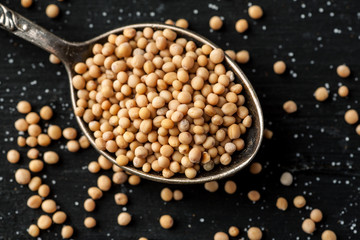 White mustard seeds in metal spoon on a black wooden table, top view, selective focus