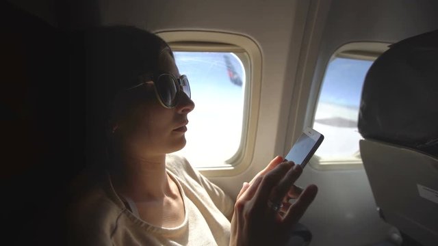 Tourist woman sitting near airplane window at sunset and using mobile phone during flight