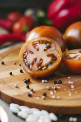 Halves of Kumato tomatoes on wooden board surrounded by vegetables and spices.