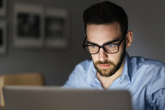 Portrait Of Handsome Bearded Man Wearing Glasses Working With Laptop In Dark Room Late At Night, His Face Lit Up By Screen