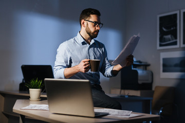 Portrait of focused bearded businessman wearing glasses working with documentation in dark room late at night, standing at workplace with heap of papers drinking coffee