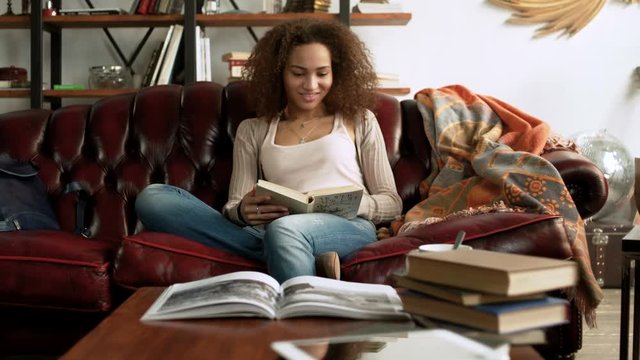 young happy Latin woman read interesting paper book for education sitting alone on comfort leather sofa in the cozy living room at home with bookshelf near window during sunny day
