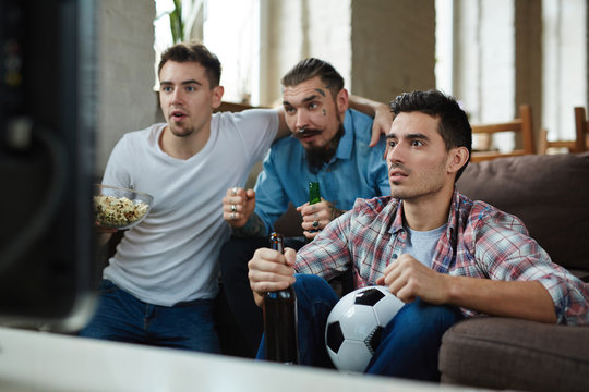 Group of friends watching football match on TV and cheering while sitting on couch and drinking beer