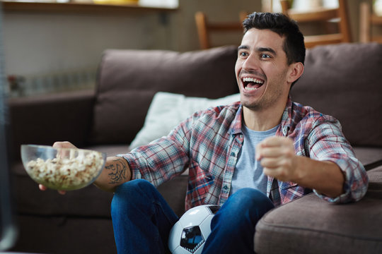 Portrait Of Excited Man Watching Sports Match On TV And Cheering Joyfully While Eating  Popcorn