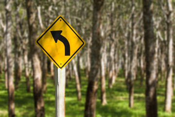 Street sign showing a bend in the road in front of rubber tree plantation in magical forest used to make latex and rubber products.