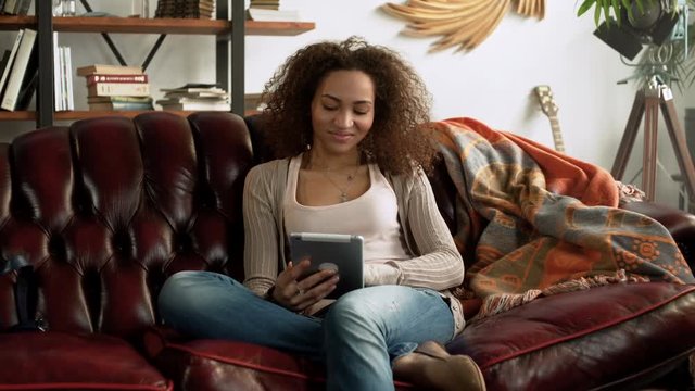young happy latin woman with curly hair use tablet computer for online communication, type messages and reading book sitting with comfort on leather sofa at home during relaxation on sunny day