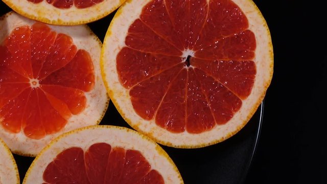 Excellent top view of fresh red grape-fruit slices on black dish, rotating clockwise with side center. Close up flat lay of yummy pieces of dessert on black background with amazing texture in 4k clip