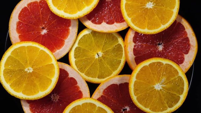 Excellent concentric top view of fresh orange and red grape-fruit slices on black dish, rotating clockwise. Flat lay of yummy pieces of dessert on black background with amazing texture in 4k clip.