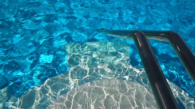 Sunlit swimming pool with clear blue water. Ripples on the water create beautiful movement. Hand rail to the water for convenient descent