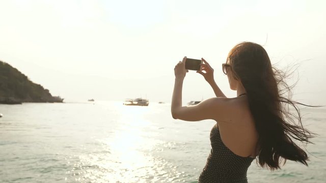 Woman tourist on beach island taking photograph of sunset with smartphone on holiday of boat and skyline view