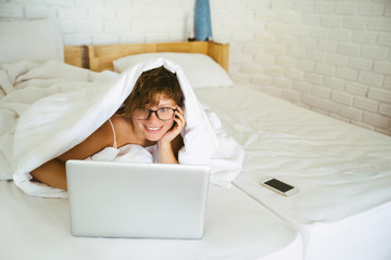 The girl in the bedroom on the bed under white blanket with a laptop in his hands. The morning starts with news on the Internet.