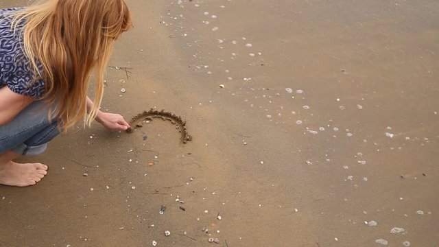 Blonde woman in love sit on the beach and draws heart symbol with his hand on the sandy floor. 