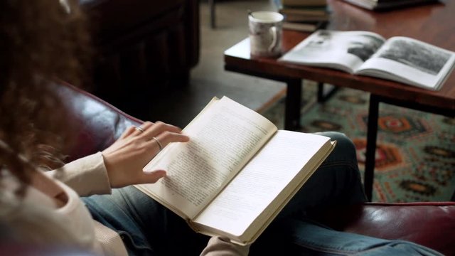 close up view of paper book in hands of latin beautiful young woman who read and studying it with interest, leaf pages with fingers, sitting on leather sofa in the room during sunny day