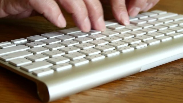 Elderly Man Is Typing On A Keyboard While Sitting At A Wooden Table.