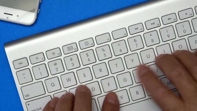 Elderly Man Is Typing On A Keyboard. On The Table Is A Mobile Phone. Blue Background.