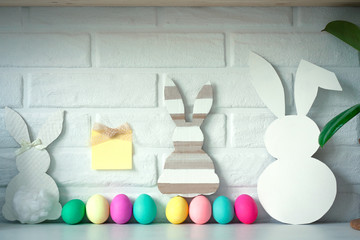 Easter white bunny with colored eggs with sticky note against white brick wall background. Cute Easter celebration decoration. Copy space for text. Happy Easter card. Easter concept.