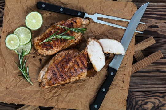 Sliced grilled chicken filet with herbs  on a paper on wooden background. Rosemary and sliced lime on a dark groundwork.