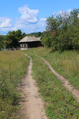 Fototapeta na wymiar Dirt road leading to a lonely wooden house in the distant Russian village against a blue cloudy sky