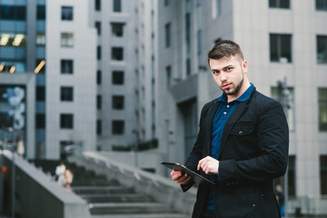 Outdoor portrait of a young handsome businessman in a suit working on a tablet on the background of a modern business center.