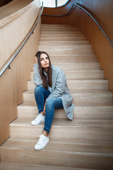 Portrait of pensive Caucasian brunette young beautiful girl woman with long dark hair and brown eyes in white shirt, grey coat and blue jeans sitting on staircase looking away