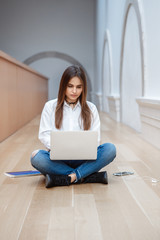 Portrait of  beautiful white Caucasian brunette young girl woman model with long dark hair in white shirt and blue jeans sitting on floor in hall at college university working on laptop