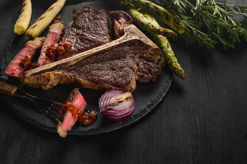Cooking steak porterhouse with vegetables on a plate and pan grill. Black table background with brown textile. Copy text area for menu design. Horizontal top view