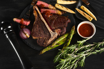 Cooking steak porterhouse with vegetables on a plate and pan grill. Black table background with brown textile. Copy text area for menu design. Horizontal top view, flat, overhead.
