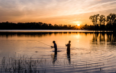Fototapeta na wymiar Two young kids are fishing under sunset of a lake