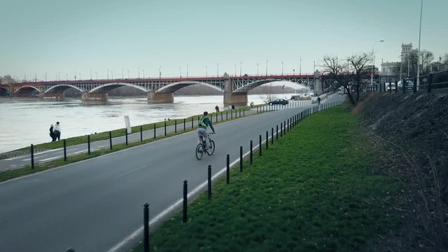 Aerial tracking shot of young man riding his bike along evening river embankment. 4K video