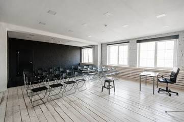 Room for lecture with a lot of dark chairs. Walls are white, loft interior. On the right there is a door. On the background there is a table with a laptop.