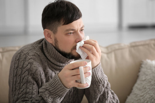 Young Ill Man Drinking Hot Tea While Sitting On Sofa At Home