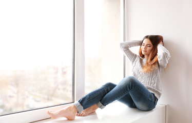 Beautiful young woman sitting on window sill at home