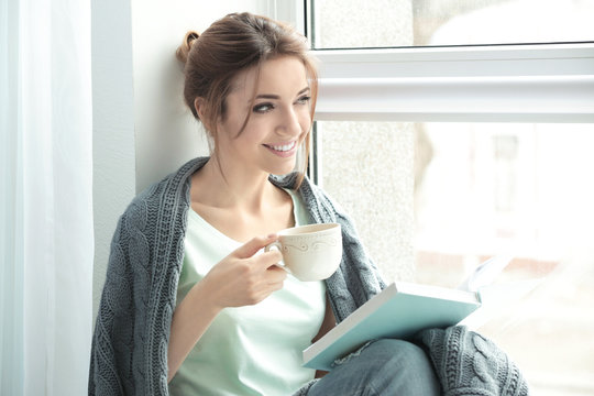 Beautiful young woman with book and cup of tea resting near window at home