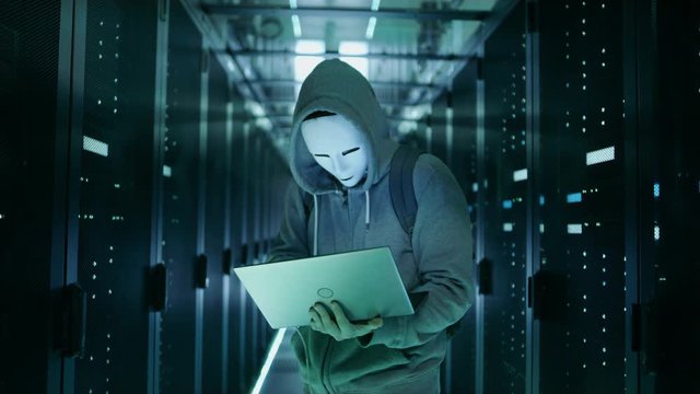 Mid Shot of a Masked Hacker in a Hoodie Standing in the Middle of Data Center full of Rack Servers and Hacking it with His Laptop.  Shot on RED EPIC-W 8K Helium Cinema Camera.