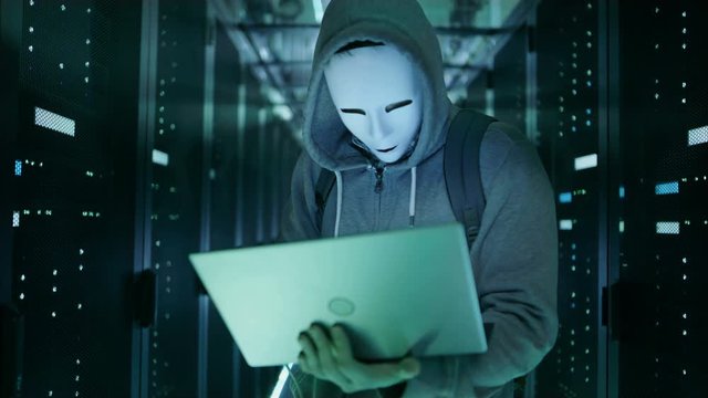 Close-up Shot of a Masked Hacker in a Hoodie Standing in the Middle of Data Center full of Rack Servers and Hacking it with His Laptop.  Shot on RED EPIC-W 8K Helium Cinema Camera.