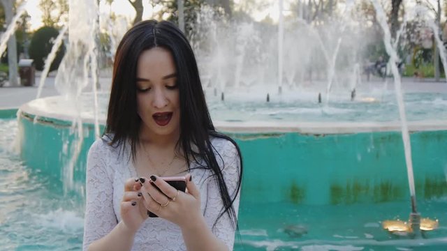 Girl pleasantly surprises using smartphone. Pretty young woman with her mobile phone on fountain background.
