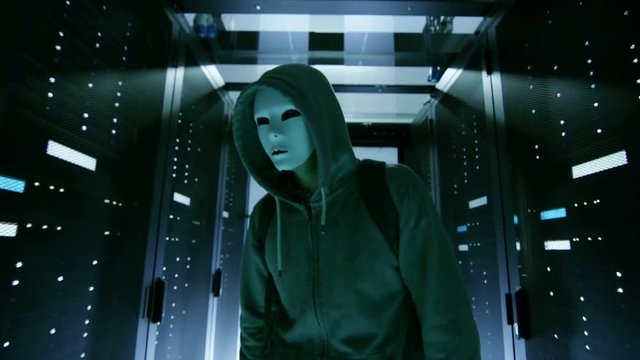 Masked Hacker in a Hoodie Walks Through Corporate Data Center with Rows of Working Rack Servers.  Shot on RED EPIC-W 8K Helium Cinema Camera.