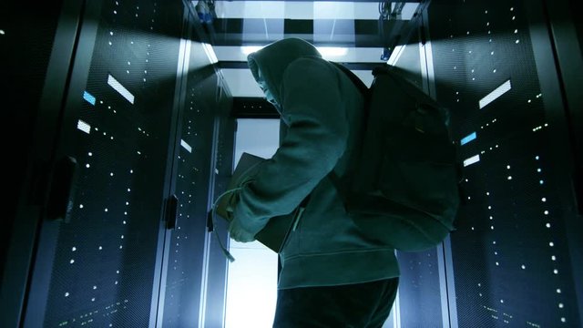 Low Angle Shot of a Masked Hacker in a Hoodie Sneaking Through Corporate Data Center with Rows of Rack Servers.  Shot on RED EPIC-W 8K Helium Cinema Camera.